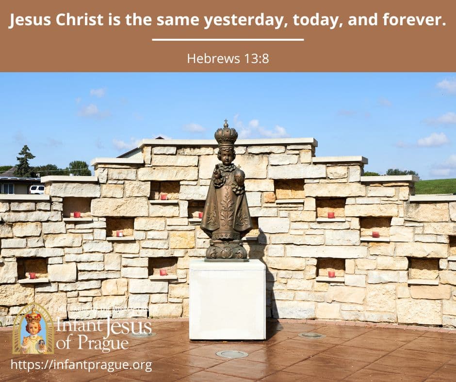 Bronze statue of the Infant Jesus of Prague in front of a stone wall with candles and a blue sky in the distance.